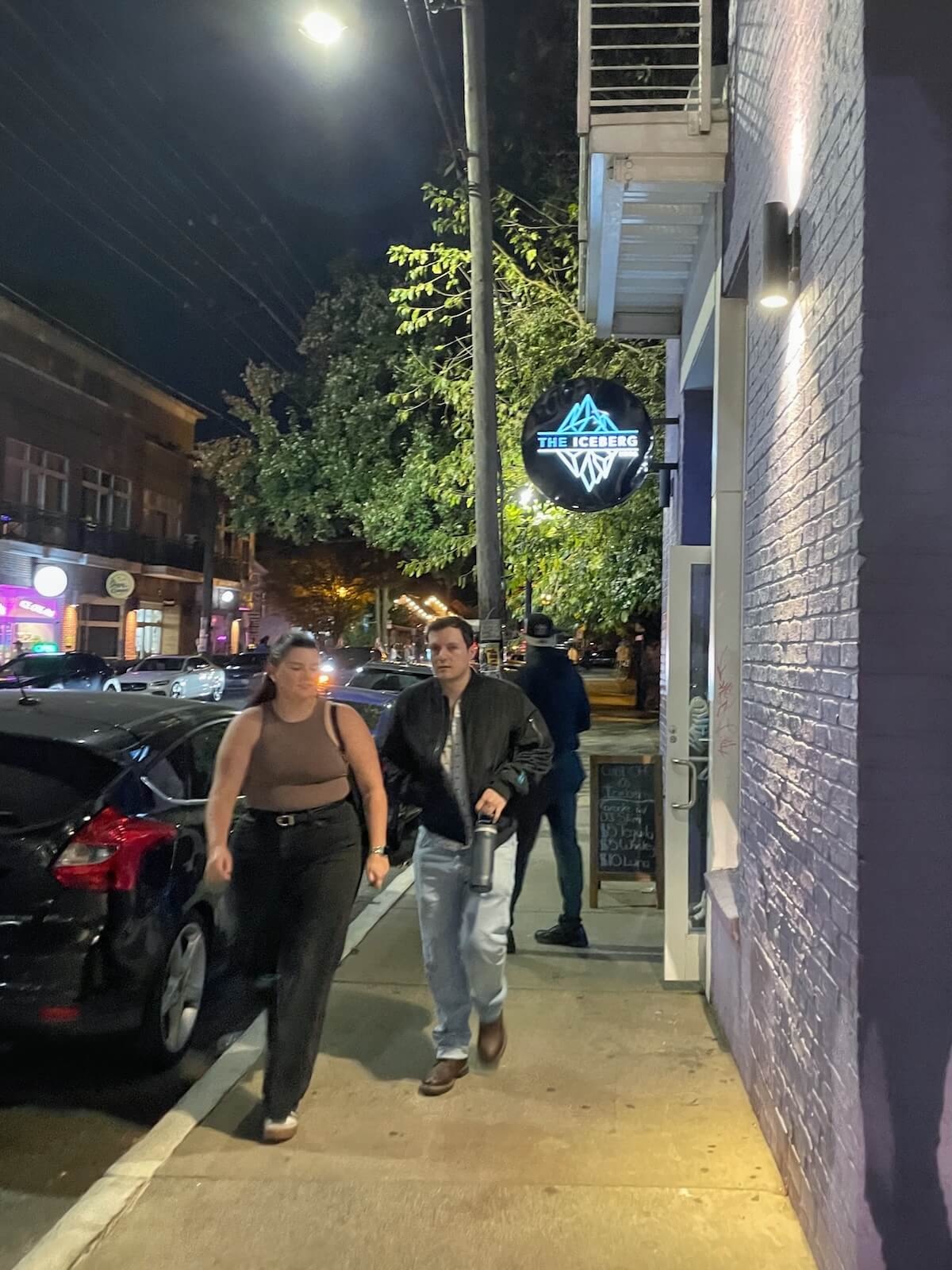 Two people walking down the street in NoDa at night. There is a building with purple wall and sign that reads Iceberg