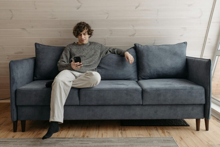 Man sitting on a blue sofa in a comfortable position while holding a cell phone.