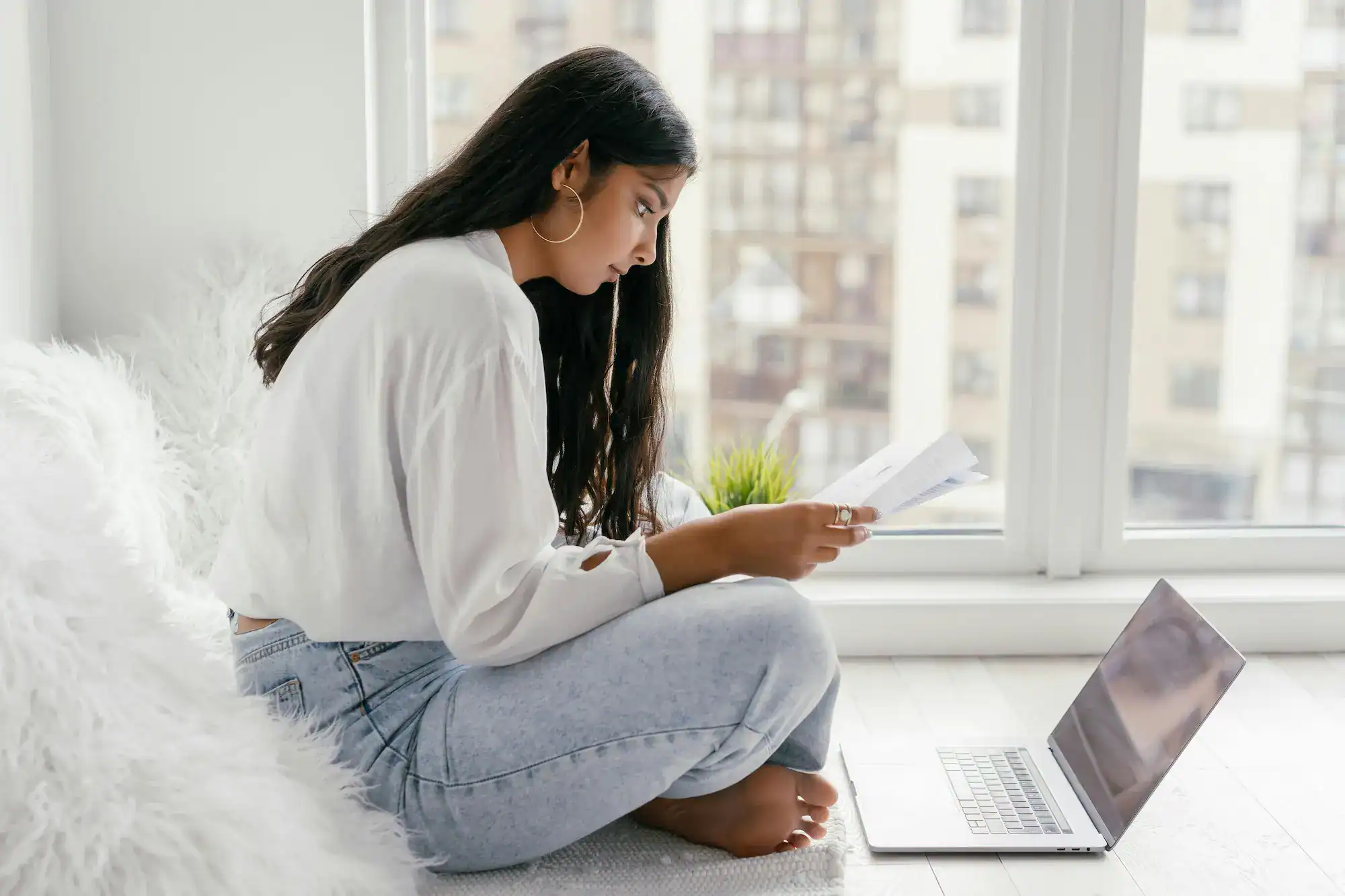 Woman sitting on the floor reading paperwork while her laptops sits open in front of her.