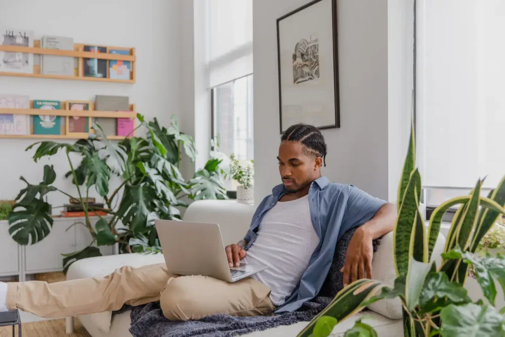 Man sitting on a sofa in a living room flooded with natural light and several plants.